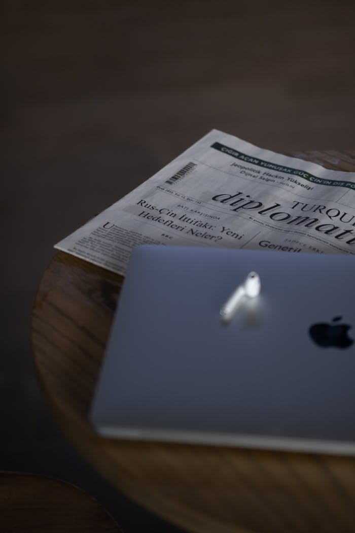 Close-up of Apple laptop with earbuds on a table, next to a folded newspaper.