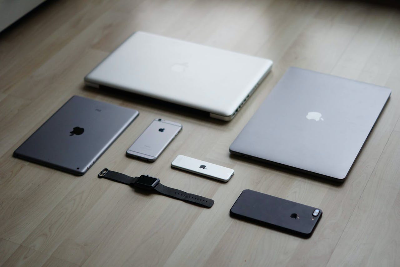 Flat lay of various Apple devices including laptops, tablets, and phones on a wooden floor.